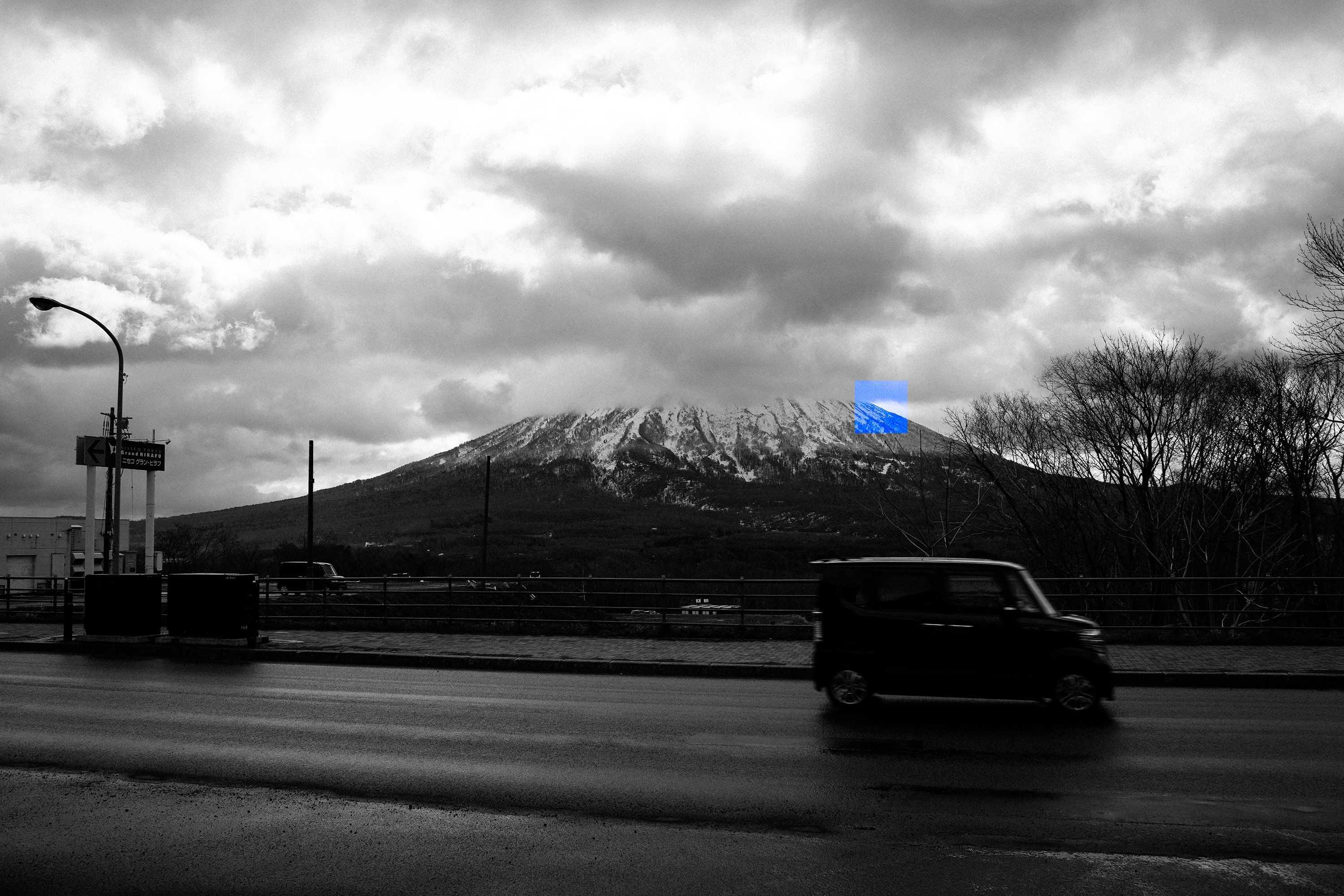 A snow-capped mountain under heavy cloud, seen across a wet road in black and white